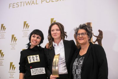 Trzy kobiety na tle ścianki Festiwalu Polskich Filmów Fabularnych w Gdyni. Kobieta w środku trzyma nagrodę Złoty Pazur