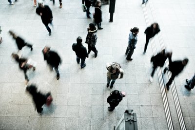 Widok z góry na chodnik, a na nim idący w róznych kierunkach ludzie, ubrani zwykle w ciemne ubrania. Niektóre sylwetki rozmazane, widać dynamikę ruchu osób i pośpiech.