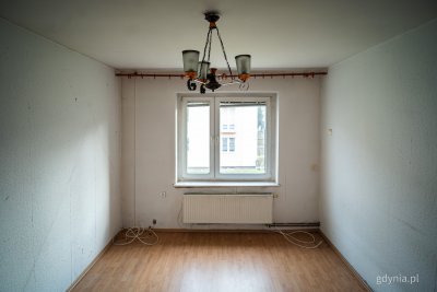 Puste pomieszczenie mieszkalne, widoczna podłoga, okno, żyrandol, grzejnik, białe ściany, do remontu