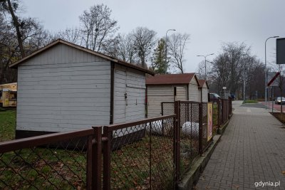 Drewniane, szare budki na trawniku, za płotem z siatki. Po prawej stronie fragment ulicy.