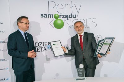 Krzysztof Jedlak i Wojciech Szczurek na tle białej ścianki. Wojciech Szczurek trzyma w rękach trzy dyplomy 