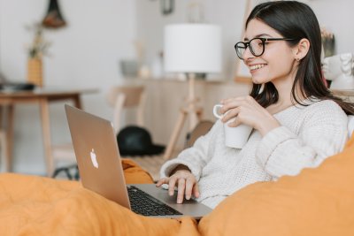 Kobieta w okularach i białym swetrze siedzi na kanapie z kubkiem i laptopem na kolanach. Jest uśmiechnięta, pracuje na laptopie
