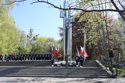 Pomnik Obrońców Wybrzeża. Na placu marynarze w mundurach i oficjele. Kilka osób składa kwiaty pod pomnikiem
