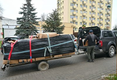 Osiedle mieszkaniowe, na parkingu bus z przyczepką, na przyczepce palety towarów. Dwie osoby układają kartony