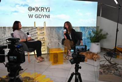 Studio do rozmów, dwie kobiety na fotelach, stolik z żółtą dekoracją, ścianka z logotypem Odkryj nieoczywiste w Gdyni, błękitne tło, sprzęt wideo
