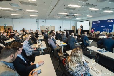 Sala konferencyjna w hotelu Marriott w Gdyni. Scena, przy mikrofonie przemawia Michał Guć, obok niego ekran z wyświetlaczem z projektora. Na sali ustawione stoły z krzesłami, przy nich siedzą ludzie w eleganckim ubiorze, światło dzienne