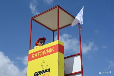 Ratownik siedzący w budce ratowniczej na plaży, obok wywieszona biała flaga.