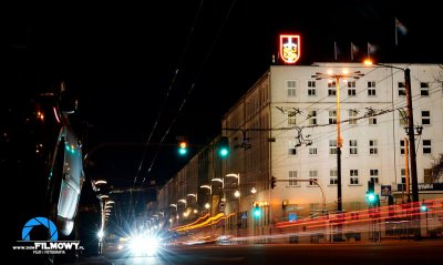 Zdjęcie skrzyżowania ul. Świętojańskiej z al. Piłsudskiego po zmroku. Widoczny budynek Urzędu Miasta Gdyni, na szczycie neonowy herb. Na poziomie ulicy rozmyte światła przejeżdżających aut