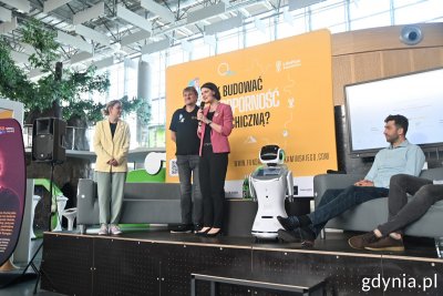 Scena w Centrum Nauki Experyment. Katarzyna Gruszecka-Spychała i Marek Kamiński przemawiają przez mikrofon. Obok nich stoi robot. Za sceną żółte tło