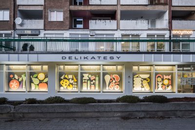 Parter bloku mieszkalnego w Gdyni. Widoczna witryna i elewacja delikatesów Aldek, jasnoszara, z elementami graficznymi i napisami
