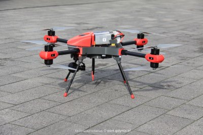 Na szarym chodniku stoi czarno-czerwony dron firmy PELIXAR. Dron posiada cztery śmigła.