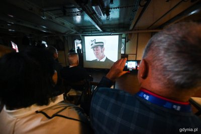 Pokłąd Daru Pomorza. Na ekranie film z marynarzem w czapce, na pierwszym planie dwie osoby oglądają film, jedna robi zdjęcie telefonem