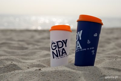 Biały i granatowy kubek z logotypem Gdyni i pomarańczową pokrywką na plaży, w piasku. W tle widać morze