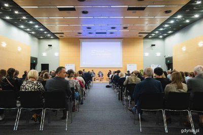 Sala konferencyjna, na środku scena i ludzie siedzący w fotelach, na krzesłach na sali widownia