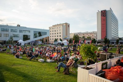 Plac Grunwaldzki w Gdyni. Na pierwszym planie na zielonej trawie leżaki i publiczność podczas festiwalu Miasto Słowa. W drugim planie biała bryła budynku Gdyńskiego Centrum Filmowego, a także Hotelu Mercure w Gdyni. Zdjęcie wykonane w dziennym, ciepłym świetle.