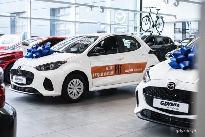 Wnętrze salonu samochodowego BMG w Gdyni, widoczne samochody na ekspozycji, przeszklona ściana, na środku stoi biała Mazda 2 z niebieską kokardą na masce, na boku naklejka z napisem 