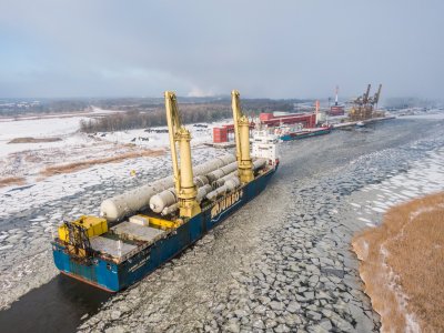 Zamarznięta rzeka, na niej statek z ładunkiem wielkogabarytowym. Na lewym brzegu rzeki widoczna przystań