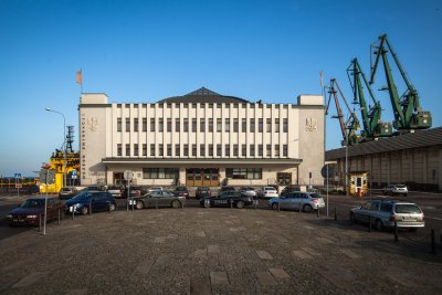 Na zdjęciu charakterystyczna bryła budynku byłego Dworca Morskiego w Gdyni, a obecnie Muzeum Emigracji w Gdyni. Po prawej stronie w tle zielone żurawie portowe. Przed budynkiem zaparkowane samochody.