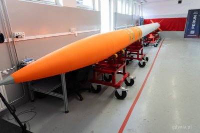 Wnętrze hali warsztatowej, biało-pomarańczowa rakieta na podporach