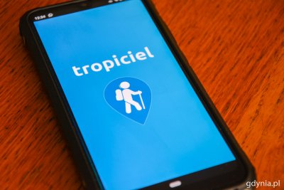 Leżący na stole Smartfon, na którego ekranie widnieje powitalny ekran aplikacji Tropiciel. Ekran jest błękitny, ma biały napis 
