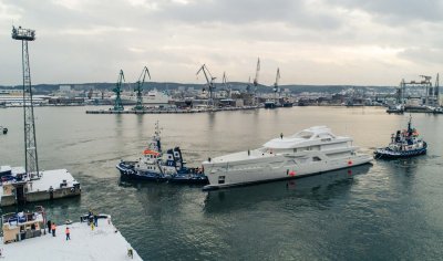 Widoczny wewnętrzny akwen portu Gdynia. Holowniki ciągną kadłub białego jachtu. W tle, na nabrzeżu widoczne stocznie