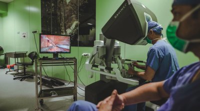 Sala operacyjna, szpital w Redłowie, kilku lekarzy w zielonych strojach, pacjent na stole operacyjnym, robot przy pacjencie, lekarz siedzi za aparaturą sterującą, wyświetla się na ekranie podgląd operacji