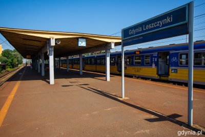 Widoczny peron kolejowy stacji SKM Gdynia Leszczynki. Brązowa nawierzchnia peronu i stara wiata przystankowa. Po prawej stronie widoczny znak z nazwą stacji. W tle zółto-niebieski pociąg SKM.