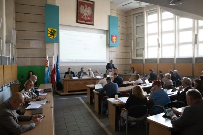 Sala obrad Rady Miasta w Urzędzie Miasta Gdyni, radni siedzą w ławach, wyświetlacz na środku, flagi, godła