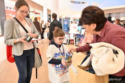 Kobieta z dzieckiem - wolontariuszem odbierają pieniądze do puszki WOŚP od starszej kobiety. Centrum Riviera, w tle hol i sklepy