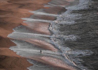 Fotografia Małgorzaty Szury-Piwnik. Brzeg morza w Portugalii, fale sięgające daleko w brzeg, widoczny mężczyzna na styku lądu i wody