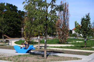 Park na Karwinach. Widoczne ścieżki w parku, ulica, drzewa i krzewy. Na pierwszym planie siedzą dwie dziewczynki odwrócone plecami, siedzą na niebieskiej ławce z tworzywa w kształcie delfina. Okres letni.