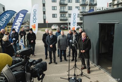 Obiekt techniczny ciepłowni - mały, szary budynek z otwartymi drzwiami, w tle osiedla mieszkaniowe, białe bloki i ulica. Przed budynkiem stoi grupa ludzi, konferencja prasowa. Kilka osób przemawia do mikrofonów na statywach, widoczni operatorzy kamer i dziennikarze, flagi promocyjne gmin