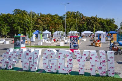 Na zdjęciu na pierwszym planie ustawiony na trawniku trójwymiarowy napis #wGdyni w drugim planie plac przy ul. Zawiszy Czarnego, na którym stoją różne, kolorowe plansze z informacjami, stoiska oraz siedziska w kształcie ryb.