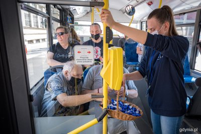 Wnętrze trolejbusu. Dziewczyna w bluzie ZKM Gdynia trzyma kosz z krówkami. Dwóch pasażerów częstuje się cukierkami. Za nimi siedzi kobieta oraz mężczyzna. W tle kilku pasażerów