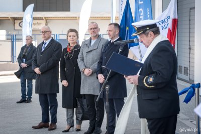 Grupa osób stoi w rzędzie, w tle flagi i budynek-hala.