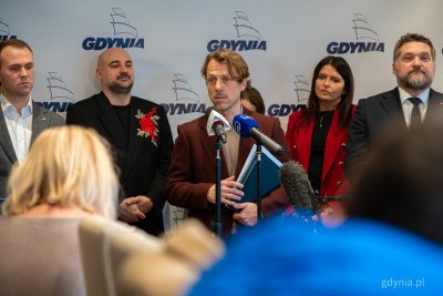 Sala w Urzędzie Miasta Gdyni, grupa radnych, na środku do mikrofonów przemawia Łukasz Piesiewicz, w tle logotypy Gdyni