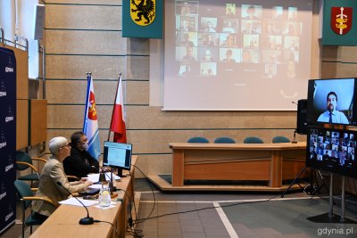 Na zdjęciu sala obrad Rady Miasta Gdyni. Po lewej stronie za stołem siedzi przewodnicząca Joanna Zielińska. Po prawej stronie ekran z radnym Tadeuszem Szemiotem, który przemawia obecnie w ramach obrad. W tle na ekranie na ścianie widoczni radni połączeni on-line.