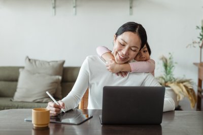 Kobieta siedzi przy stole i pracuje na laptopie. Na szyi opiera się jej dziecko, uśmiecha się. W tle mieszkanie