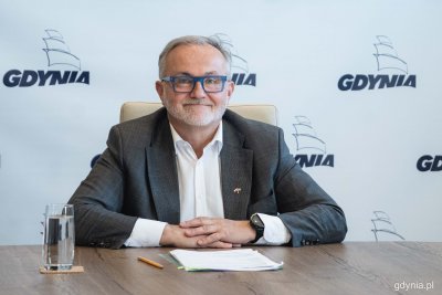 Wojciech Szczurek siedzi przy stole. Patrzy w obiektyw, uśmiecha się, jest ubrany w garnitur. Za nim biała ściana z logotypami Gdyni