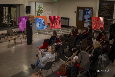 Kobieta stoi i przemawia do zebranej publiczności, w tle na sztalugach znajdują się trzy wielkoformatowe ilustracje gdyńskiej artyski.