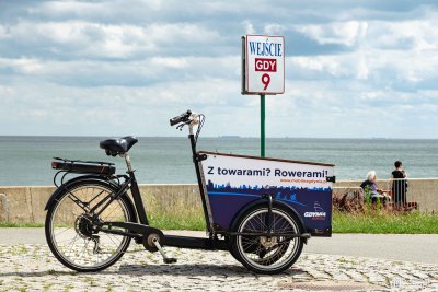 Rower cargo ze skrzynią w biało-niebieskim kolorze, stoi na kostce brukowej na bulwarze Nadmorskim w Gdyni, w tle betonowy falochron, morze, niebo, słoneczny dzień