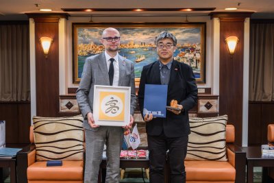Sala w budynku należącym do miasta Kaohsiung. W tle dekoracje, obraz z nadmorskim miastem, na środku pozują do zdjęcia Bartłomiej Austen i Lin Rongbin, trzymają sprezentowaną grafikę oraz teczkę