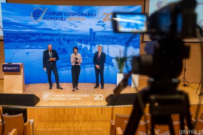 Kamera na pierwszym planie. Na drugim planie scena, na scenie trzy osoby przemawiają przez mikrofon. Widoczna dekoracja z logo Forum Gospodarki Morskiej Gdynia.