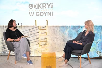 Studio Gdynia Filmowa, dwie kobiety siedzą w fotelach i rozmawiają, na środku stolik z żółtą dekoracją, w tle plansza z błękitnym tłem i napisem 