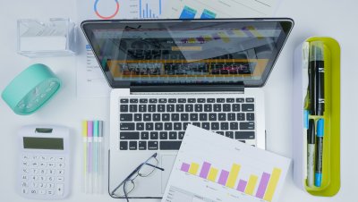 Otwarty laptop na stole, obok długopisy, wykresy na kartkach, przybory piśmiennicze