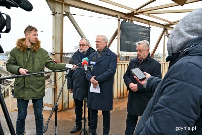 Kładka dla pieszych na SKM Gdynia Stocznia. Troje mężczyzn przemawia przy mikrofonach, obok dziennikarze i kamery