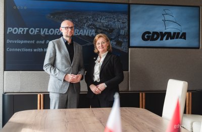 Sala konferencyjna w Urzędzie Miasta Gdyni, wiceprezydent Austen pozuje do zdjęcia z ambasador Mimozą Halimi. Za nimi dwa wyświetlacze ze zdjęciami portu we Wlorze oraz logotypem Gdyni.