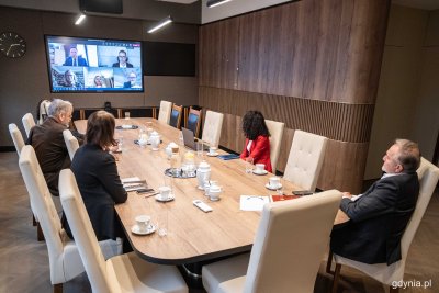 Sala, duży stół, przy stole kilka osób, spoglądają w stronę ekranu. Na ekranie kilka osób na łączeniu online