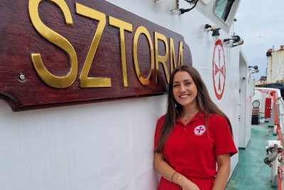Pokład statku ratowniczego, napis Sztorm na burcie, do zdjęcia pozuje młoda kobieta, brunetka w czerwonym uniformie ratowników morskich