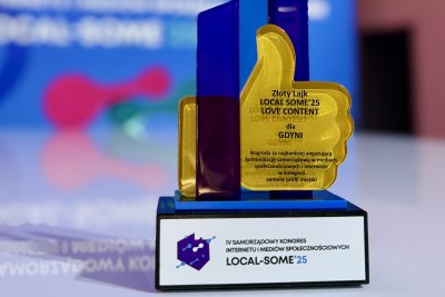 Ekspozycja, białe tło, logotyp kongresu Local Some, na pierwszym planie statuetka ze złotym, uniesionym kciukiem